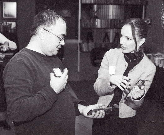 Two members talking at a club event, in black and white: a man in glasses holding a drink and a phone, a woman in a cardigan and turtleneck gesturing as she speaks, bookshelves behind them.