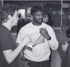 Two founders talking at a club evening, one in a white sweatshirt holding a drink, the other listening