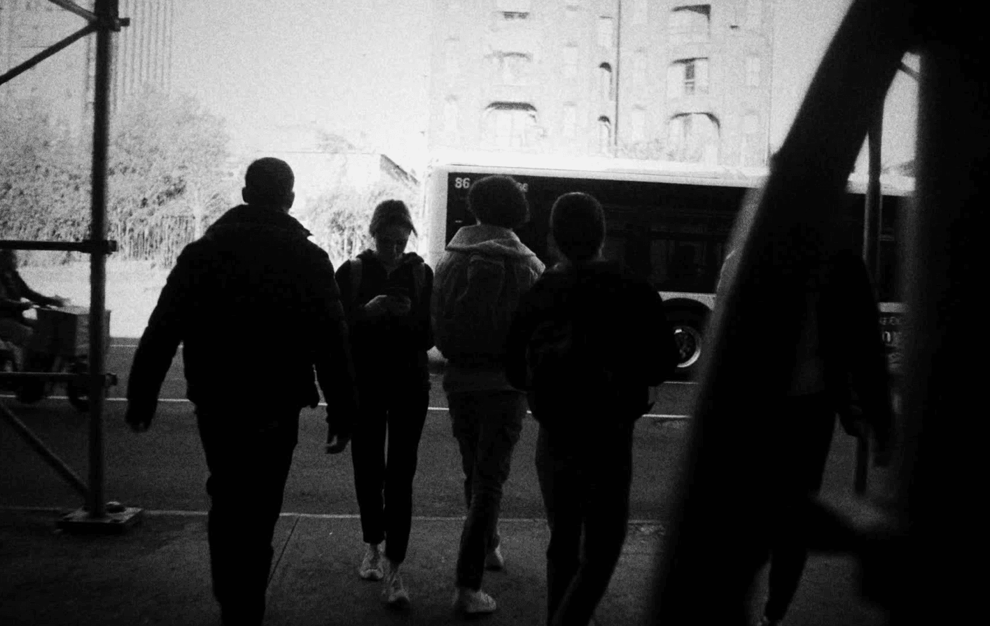 A group of people in dark coats walk out from under a scaffolding shed onto a New York street as a city bus pulls past.