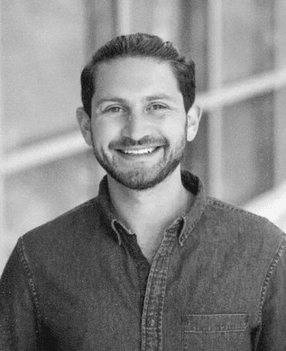 Barak Kaufman with a short beard, smiling, in a denim shirt in front of a window